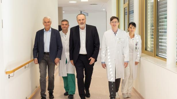 Fünf Personen, darunter Ärzte in weißen Kitteln und zwei Männer in Anzügen, gehen gemeinsam durch einen hellen Krankenhausflur.