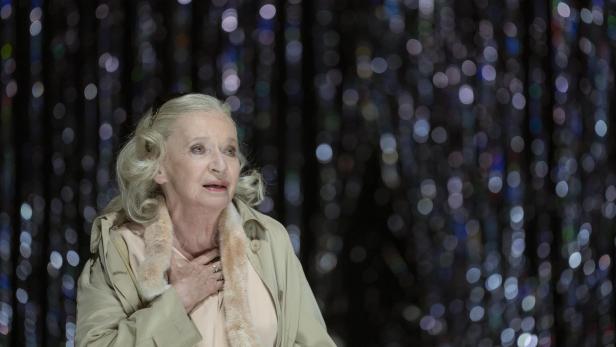 Ältere Frau mit blondem Haar und beigem Mantel hält sich die Hand an die Brust vor einem glitzernden, unscharfen Hintergrund.