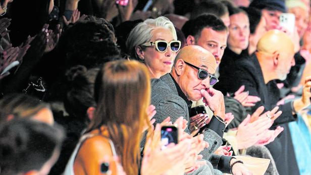 Zuschauer applaudieren bei einer Modenschau, darunter eine Frau mit weißem Haar und ein Mann mit Sonnenbrille.