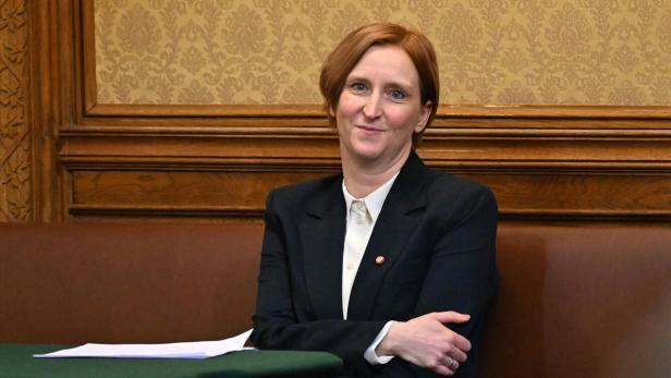 Elke Hanel-Torsch mit kurzem, rötlichem Haar sitzt in einem eleganten Raum auf einer Bank und trägt einen dunklen Blazer.