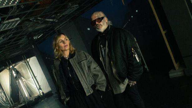 Zwei Personen stehen in einem dunklen Raum mit offenem Deckengebälk, über ihnen hängt eine Schaufensterpuppe kopfüber.