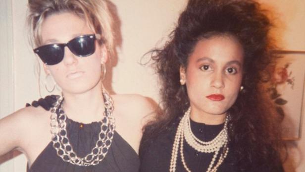 Zwei Frauen mit ernstem Gesichtsausdruck und auffälligem 80er-Jahre-Stil posieren nebeneinander.