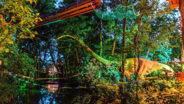 Das Lebensgroße Modell vom Brachiosaurus ist mit bunten Lichter beleuchtet und steht im Gebüsch.