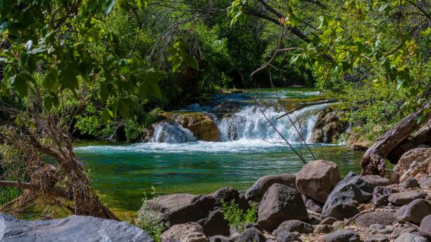 Kleiner Wasserfall mit klarem Wasser, umgeben von Felsen und dichter grüner Vegetation.