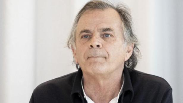 Ein Mann mit grauen Haaren sitzt an einem Tisch mit Namensschild, Wasserflasche, Glas und Mikrofon vor hellem Hintergrund.