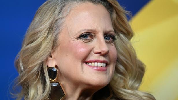 Barbara Schöneberger mit blonden, gewellten Haaren und auffälligen Ohrringen vor farbigem Hintergrund.