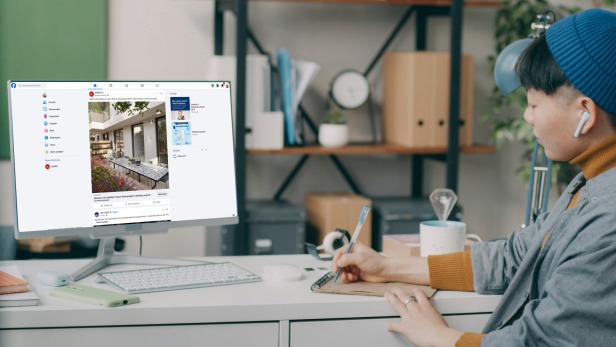 Person mit Mütze und Ohrhörern sitzt an Schreibtisch und schreibt neben einem Monitor mit geöffneter Social-Media-Seite