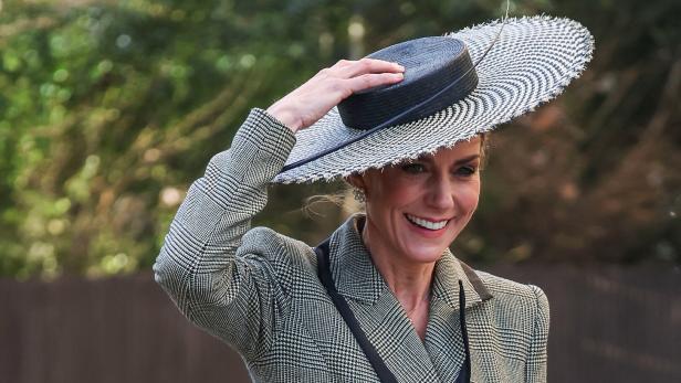 Prinzessin Kate hält ihren Hut mit breiter Krempe und lächelt in einem karierten Blazer.