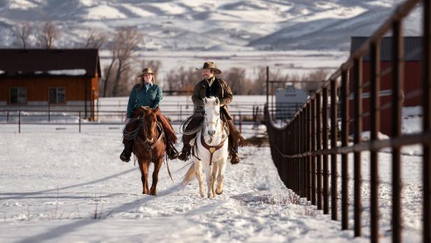 Zwei Reiter reiten auf verschneitem Reitplatz neben einem Zaun, im Hintergrund Berge und Stallgebäude.