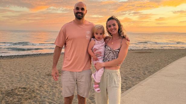 Gus und Emily Forrester stehen mit ihrer Tochter Leni am Strand und blicken gemeinsam in die Kamera, während die Sonne untergeht.