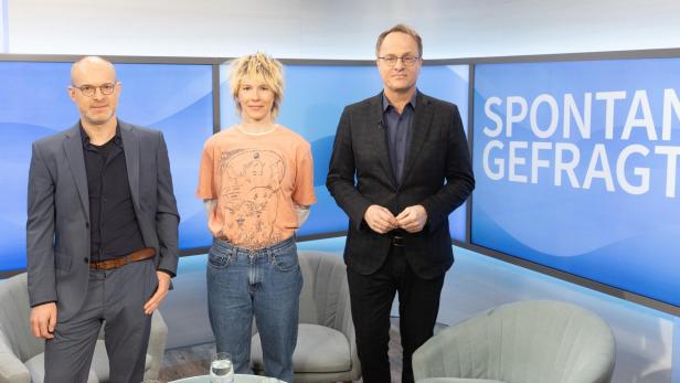 Drei Personen stehen vor einem blauen Hintergrund mit dem Schriftzug 'SPONTAN GEFRAGT', zwei tragen Anzüge, eine Person trägt ein T-Shirt und Jeans