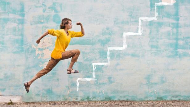 Eine Frau springt dynamisch vor einer Wand mit aufgemalten weißen Treppenstufen nach oben.