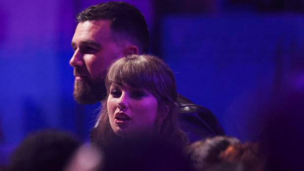 Taylor Swift und Travis Kelce stehen nebeneinander in einer Menschenmenge bei violettem Licht.