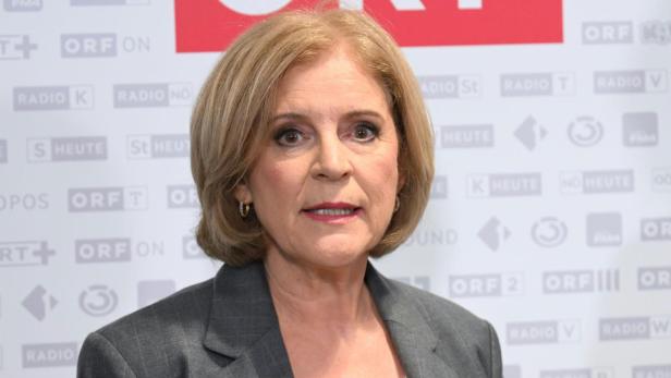 Eine Frau mit blondem, schulterlangem Haar trägt einen grauen Blazer und steht vor einer Wand mit ORF-Logos.