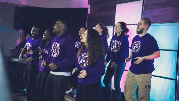 Mehrere Menschen singen und tanzen gemeinsam auf einer Bühne in lila Pullovern.