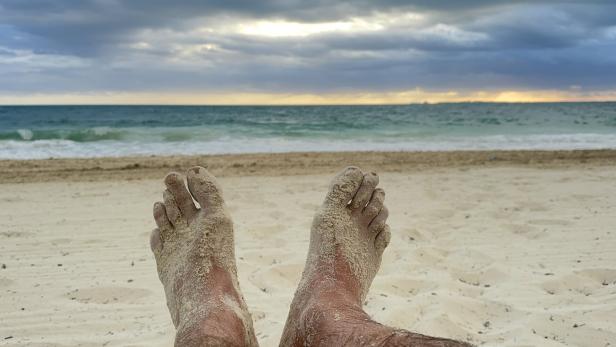 Sandige Füße entspannen am Strand mit Blick auf das Meer unter bewölktem Himmel.