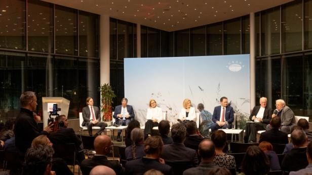 Podiumsdiskussion mit mehreren Teilnehmern vor Publikum im modern beleuchteten Saal.