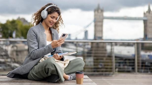 Junge Frau mit Kopfhörern sitzt draußen vor der Tower Bridge, schaut lächelnd auf ihr Smartphone und hält Notizbücher.