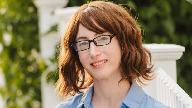 Eine Person mit braunen, welligen Haaren und Brille trägt ein hellblaues Hemd und steht vor einem weißen Geländer im Grünen.