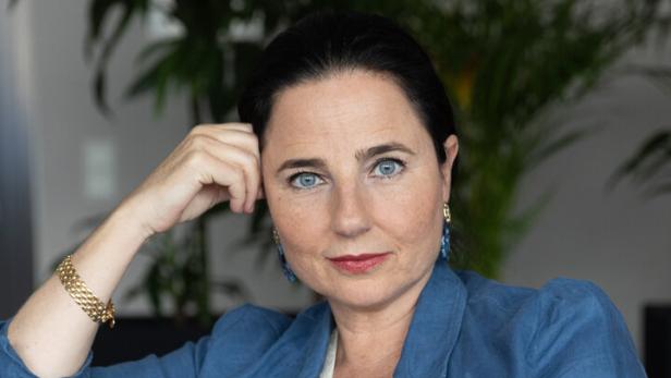 Frau mit dunklen Haaren und blauen Augen sitzt auf einem roten Sofa, trägt einen blauen Blazer und schaut direkt nach vorn.