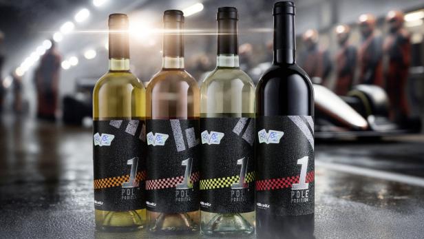 Vier Weinflaschen mit Rennsport-Etiketten stehen nebeneinander in einer Boxengasse mit unscharfen Personen im Hintergrund.