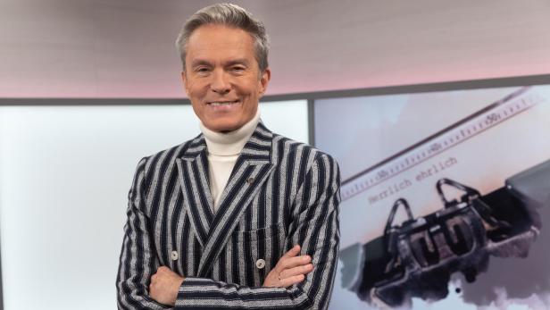 Ein lächelnder Mann mit gestreiftem Sakko und Rollkragenpullover steht vor einem Bildschirm.