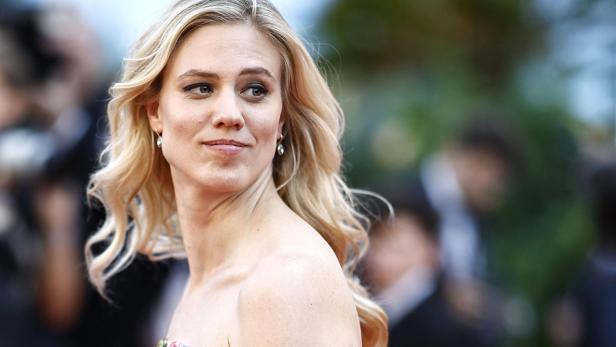 Eine Frau mit langen blonden Haaren und schulterfreiem Kleid blickt zur Seite, im Hintergrund unscharfe Menschen.