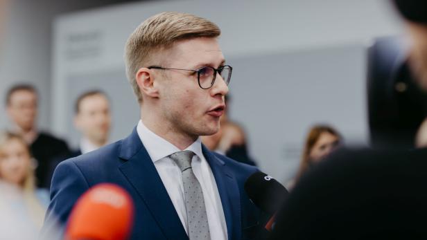 Ein Mann mit Brille und Anzug spricht in Mikrofone vor unscharfem Hintergrund.