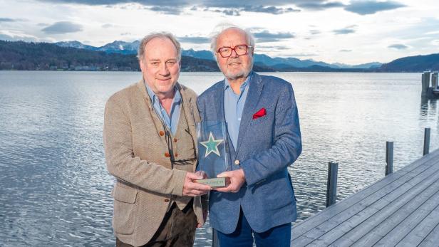 Zwei Männer halten eine Auszeichnung vor einem See und Bergen.