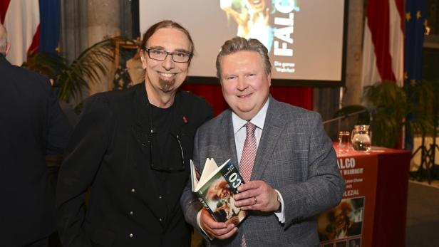 Zwei Männer posieren mit einem Buch über Falco, im Hintergrund eine Leinwand mit dem Falco-Schriftzug.