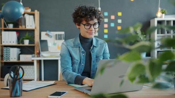 Person mit lockigem Haar und Jeansjacke sitzt an einem Schreibtisch und arbeitet an einem Laptop