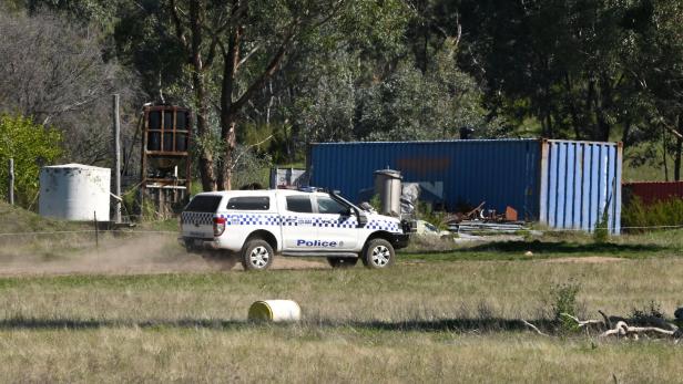 Ein Polizeiauto fährt auf einem Feldweg an einem blauen Container und verschiedenen Metallgegenständen vorbei.