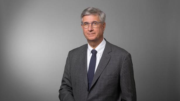 Ein älterer Mann mit grauen Haaren, Brille und Anzug steht vor einem grauen Hintergrund und blickt freundlich nach vorn.