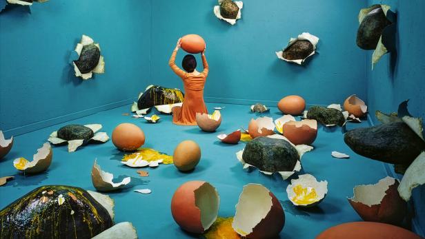 Eine Person in orange sitzt in einem blauen Raum voller zerbrochener Rieseneier und Steine, die aus den Wänden ragen.