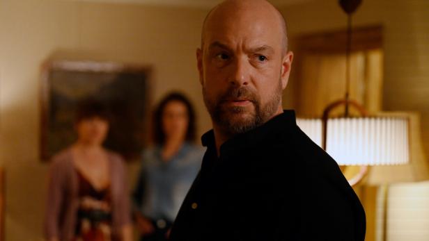 Pfarrer Reiser (Stephan Zinner) mit Bart und schwarzem Hemd blickt aufmerksam, während zwei Frauen im Hintergrund stehen.