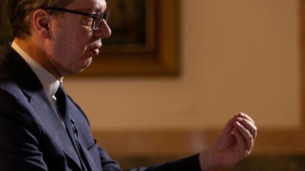 Ein Mann mit Brille und Anzug gestikuliert vor einem beigen Hintergrund.