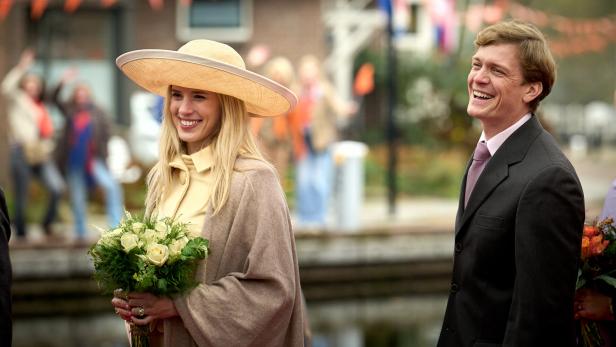 Máxima mit Hut hält einen Blumenstrauß, neben ihr ein lachender Willem-Alexander im Anzug.