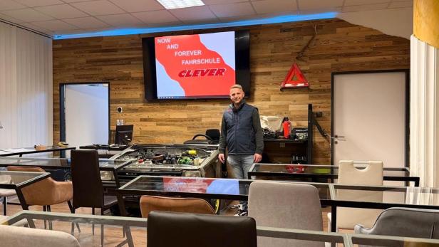 Ein Fahrlehrer der Fahrschule Clever Graz steht in einem modernen Unterrichtsraum mit Motorenmodell und Präsentationsbildschirm.
