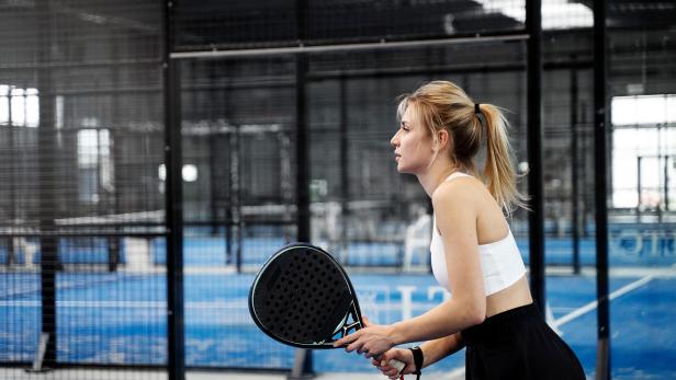 Junge Frau mit blondem Pferdeschwanz hält einen Padel-Schläger und steht konzentriert auf einem blauen Padelplatz.