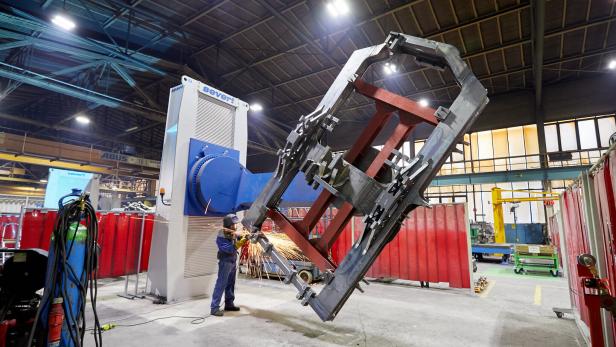Eine Person schweißt an einem großen Metallrahmen in einer Industriehalle mit moderner Maschinenanlage.