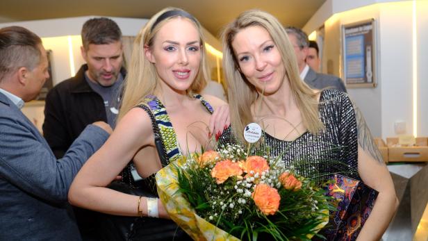 Zwei Frauen lächeln und halten einen Strauß orangefarbener Blumen, im Hintergrund stehen mehrere Personen.