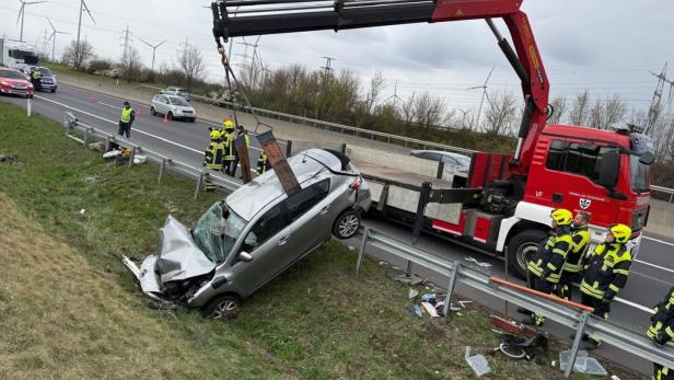 Ein silbernes Auto mit starkem Frontschaden wird von Feuerwehrleuten und einem Kran von einer Böschung neben einer Straße geborgen.