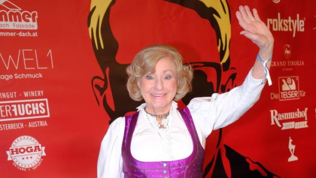 Margot Hellwig im lila Dirndl vor rotem Hintergund.