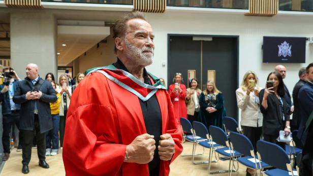 Arnold Schwarzenegger trägt eine rote Robe und blickt in die Ferne, während Leute Fotos machen.