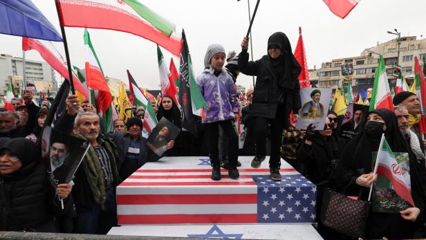 Zwei Kinder stehen auf einer Plattform mit US-Flagge und Davidstern, umgeben von Menschen mit iranischen Fahnen und Porträts.