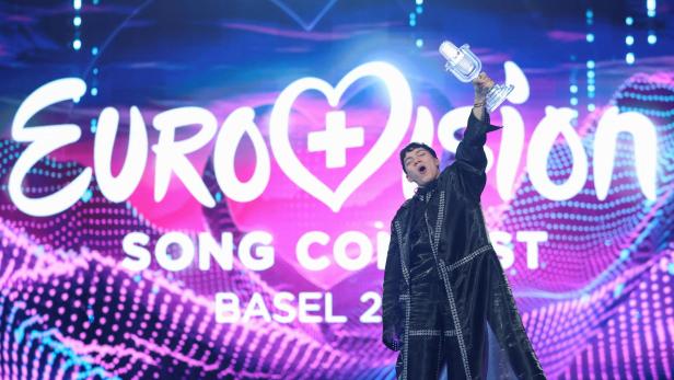 Eine Person hält jubelnd einen gläsernen Mikrofon-Pokal vor einer bunten Eurovision Song Contest Bühne hoch.