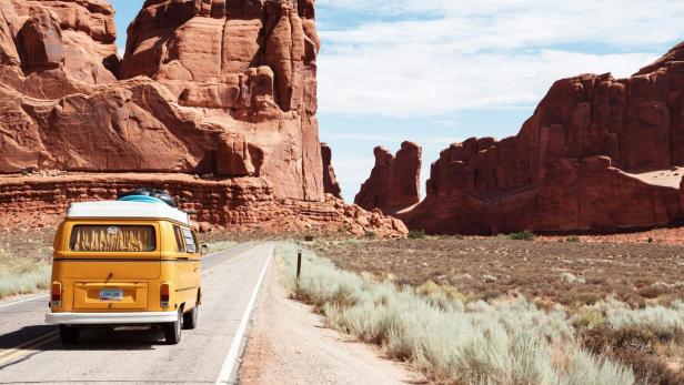 Gelber Campingbus fährt auf Straße durch beeindruckende rote Felslandschaft bei sonnigem Wetter.