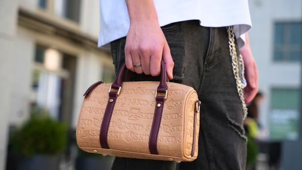 Beige SKD-Handtasche mit geprägtem Logo und braunen Henkeln wird von einer Person getragen.