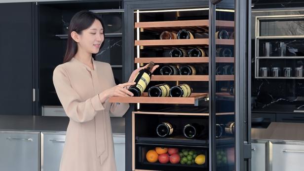 Eine Frau stellt eine Weinflasche in den offenen Samsung KI-Weinkühlschrank, der mit Holzregalen und einem Fach für Obst ausgestattet ist.