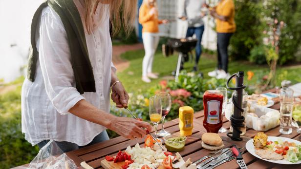 Frau bereitet Gemüse am Tisch vor, im Hintergrund grillen mehrere Personen im Garten.